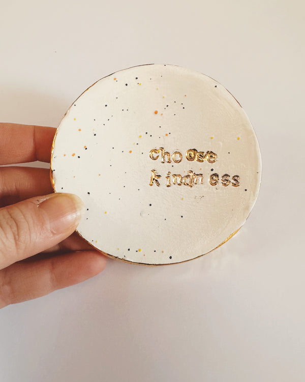 Round speckled ceramic dish with 'Choose Kindness' text held by a hand on a light background