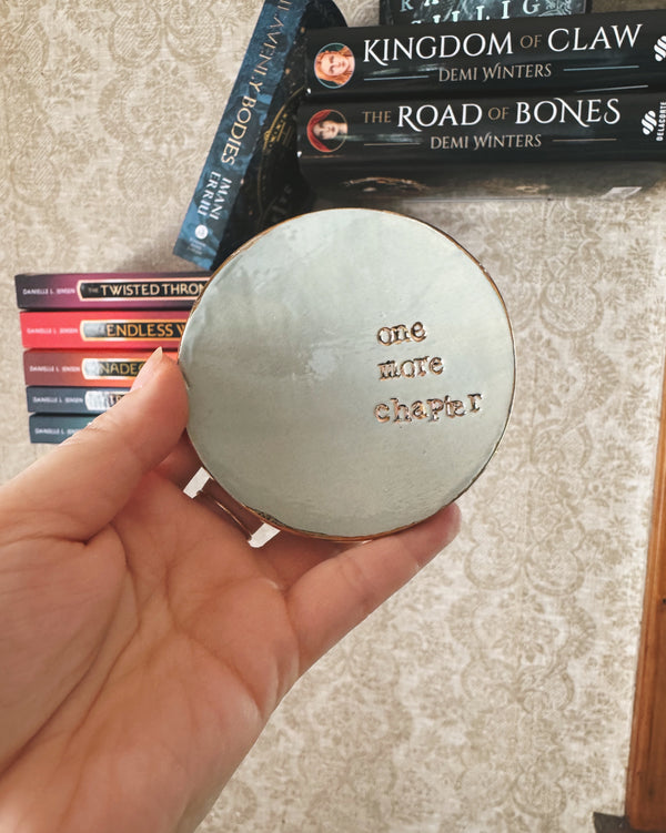 Round ceramic object with 'One More Chapter' engraved, held over books on a textured surface